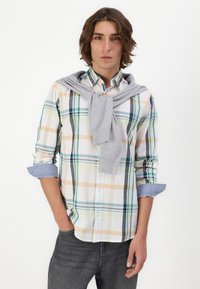 Chemise à carreaux blanche, bleue et beige avec les manches retroussées. Un pull gris drapé sur les épaules. Jean foncé.