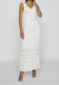 Vit, ärmlös maxiklänning med V-ringning, strukturerad tyg, horisontella veck och utsvängd nederkant. Kombineras med sandaler med remmar och dekorationer.