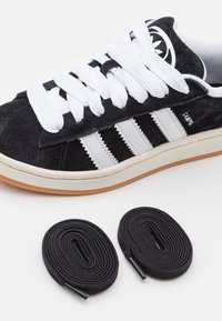 Sneaker en suède noir avec trois rayures blanches, lacets ronds blancs et semelle en gomme ; comprend deux lacets noirs avec embouts en plastique.