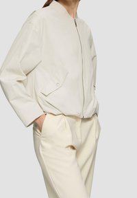 Light beige cropped jacket with a zip front, stand collar, and a side pocket. Worn with matching light beige tailored pants.