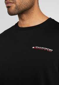 Černé bavlněné tričko s kulatým výstřihem. Na levé straně hrudi výrazné bílo-červené logo "TOMMY SPORT", hladká textura, volný střih.