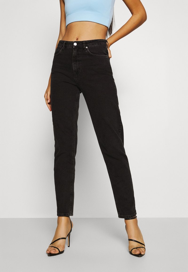 Femme portant un jean slim taille haute noir, un top court bleu clair et des sandales noires à talons hauts à brides, debout devant un fond uni.