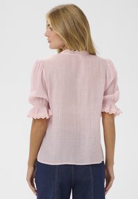 Blouse rose clair avec manches bouffantes, tissu texturé, encolure festonnée et bordure en dentelle sur les bords des manches. Portée sur un jean denim bleu foncé.
