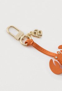 Kuldne metallist klamber graveeringuga "BOSS", kinnitatud oranžile nahast homaari võtmehoidjale, millel on kuldne "BB" ripats valgel taustal.