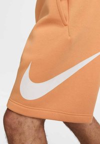 Pantalones cortos Nike naranja con un gran logo de swoosh blanco, hechos de material suave con detalles de costura visibles y bolsillos laterales.