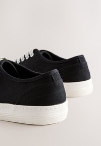 Baskets en toile noire avec semelles en caoutchouc blanches et lacets blancs, vues de dos sur un fond clair uni.