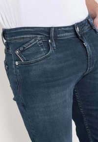 Jean en denim bleu foncé avec une coupe slim, comportant une poche avant avec une étiquette de marque et des fermetures à bouton en métal sur la taille.