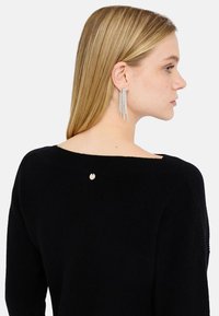 Maglione nero in maglia con collo ampio, dotato di un piccolo accento dorato sul retro e orecchini pendenti trasparenti in strass.