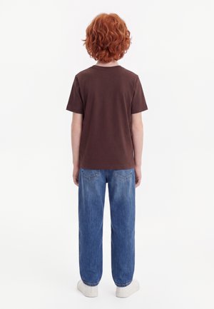 Barn med krøllet rødt hår står med ryggen mod, iført en brun kortærmet skjorte, blå jeans og hvide sneakers på en hvid baggrund.