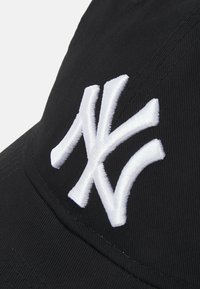 Mustmust, millel on silmapaistev valges tikandiga 'NY' logo, kaldu, et näidata kangast ja õmblusdetailide tekstuuri.