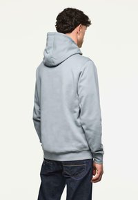 Felpa grigia con cappuccio realizzata in un mix di cotone; presenta una tasca a marsupio e polsini a coste; indossata con jeans di jeans scuri. Vista da dietro.