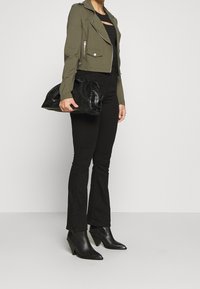 Giacca cropped verde oliva con dettagli in argento, top aderente nero, pantaloni neri a campana, stivali neri con tacco e una clutch nera testurizzata.