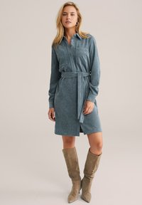Robe chemise en velours côtelé bleu avec des manches longues, une poche avant, une fermeture à boutons et une ceinture assortie, associée à des bottes taupe arrivant aux genoux.