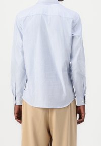 Muž v světle modré a bílé pruhované košili s dlouhým rukávem a krémových kalhotách, zobrazený zezadu na neutrálním pozadí.