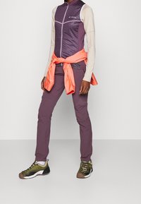 Chaleco aislante morado con patrón geométrico, camisa de manga larga crema, pantalones morados y zapatos de senderismo verdes. Acentos en naranja brillante.