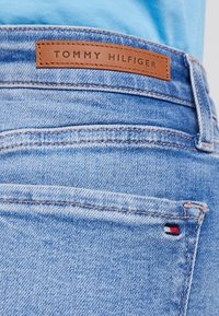 Blå denimjeans med en brungul läderetikett märkt "TOMMY HILFIGER" och en liten tri-färg logo på bakfickan. Synliga avslutningar.