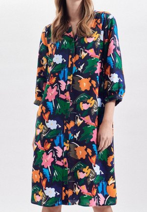 Vrouw draagt een knielange jurk met knopen en 3/4 mouwen met een kleurrijk abstract bloemenpatroon op een donkere achtergrond.