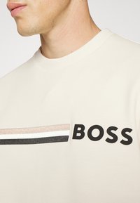 Gros plan d'une personne portant une chemise crème à rayures horizontales avec le texte noir en gras « BOSS » sur la poitrine.