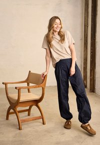 Smilende kvinde i beige skjorte og marineblå bukser står ved siden af en træarmstol med beige hynder i et minimalistisk indendørs rum.