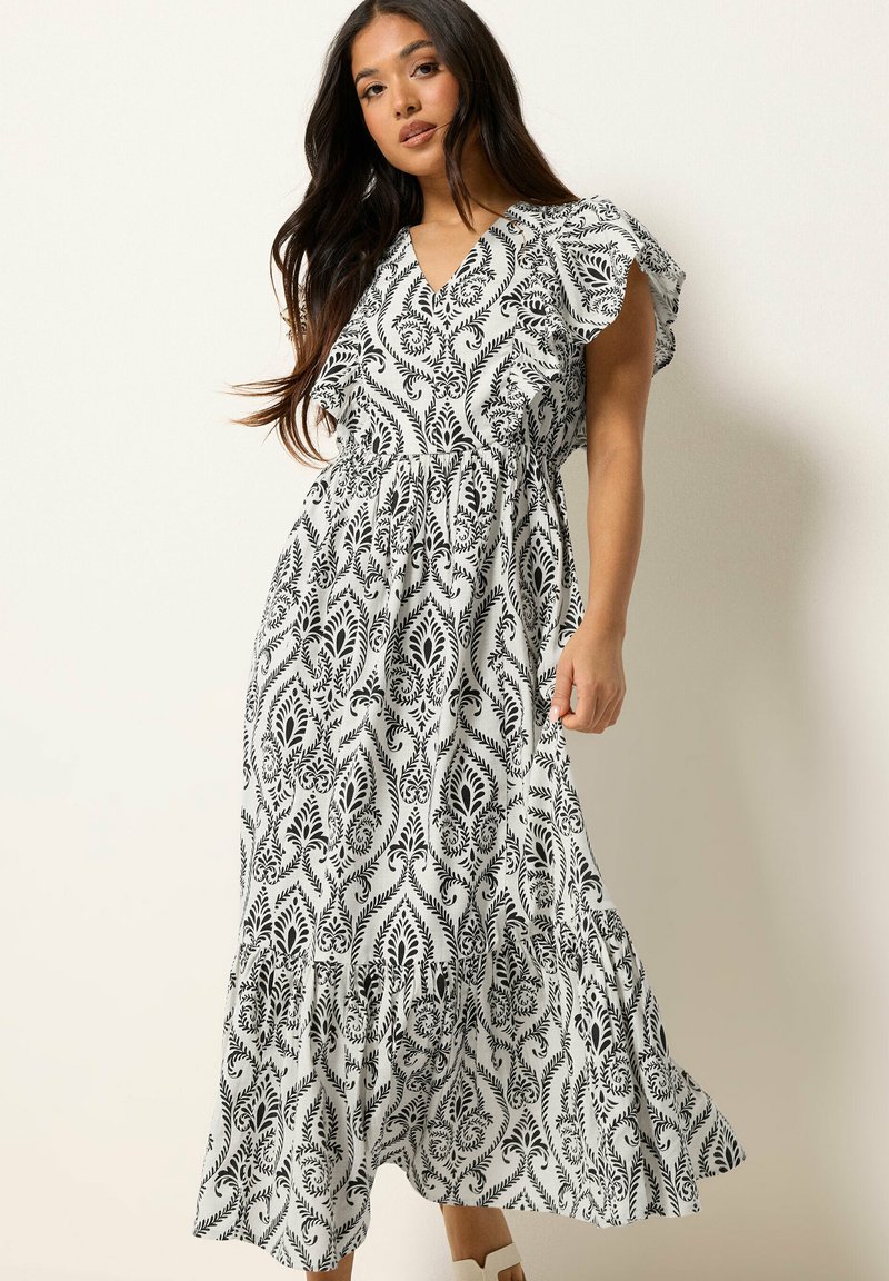 Junge Frau trägt ein schwarz-weiß gemustertes Maxikleid mit flatternden Ärmeln und steht vor einem schlichten hellen Hintergrund.