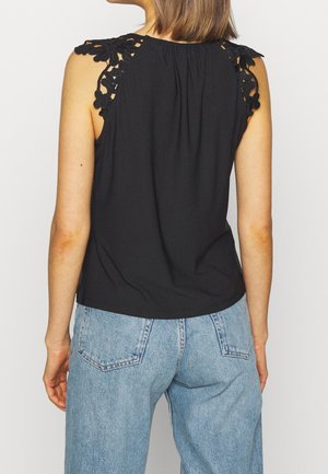 Kobieta ubrana w czarny, bezrękawnikowy top z koronkowymi kwiatowymi wstawkami na ramionach oraz jasnoniebieskie, wysokowalne dżinsy, zwrócona tyłem do kamery.