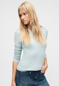 Ξανθιά γυναίκα με μακριά μαλλιά που φορά ένα ανοιχτό μπλε ριπ πουλόβερ και μπλε τζιν, κοιτάζοντας προς τα αριστερά της με το ένα χέρι στην τσέπη της.