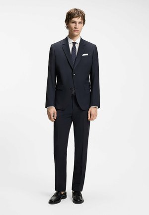 Joven vestido con un traje azul marino oscuro, camisa blanca, corbata azul marino con estampado, pañuelo blanco en el bolsillo y zapatos de cuero negros, posando sobre un fondo blanco.