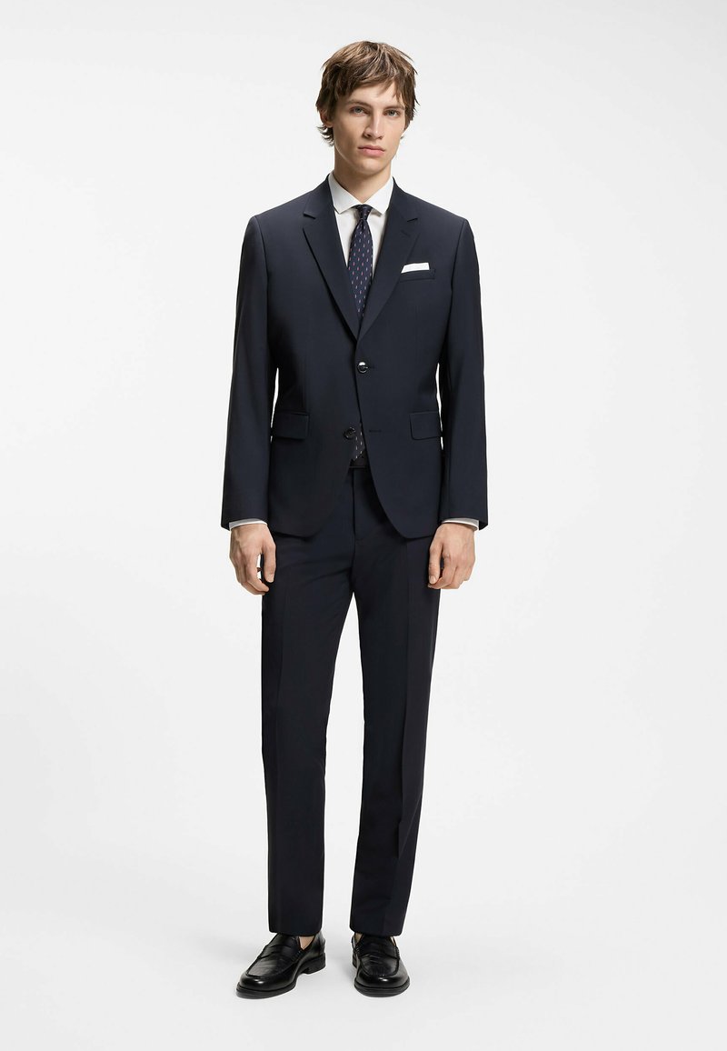 Jeune homme portant un costume bleu marine foncé avec une chemise blanche, une cravate bleu marine à motif, une pochette blanche et des chaussures en cuir noires, debout devant un fond blanc.