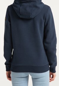 Marinblå hoodie med ett strukturerat tyg, ribbade ärmslut och fåll, samt en huva med snörning. Har liten logotyp på den övre ärmen.