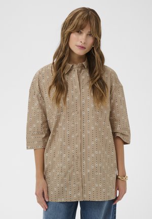 Hosszú barna hajú nő bézs színű, gombos, függőleges virágmintás csíkos ingben és kék farmerral, fehér háttér előtt állva.