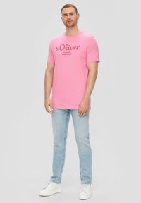 Roze katoenen t-shirt met korte mouwen en een grafische tekst aan de voorkant. Gecombineerd met lichtblauwe jeans en witte sneakers voor een casual look.