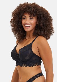 Bralette in pizzo nero con motivi floreali e bordi a scallop. Spalline regolabili e chiusura frontale decorativa. Texture morbida, design trasparente.