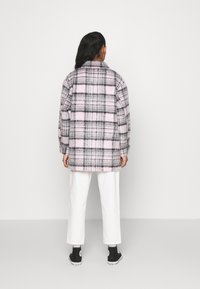Chaqueta de cuadros oversized en gris y rosa, hecha de un tejido suave con una apariencia texturizada. Llevada sobre pantalones blancos y zapatillas negras.