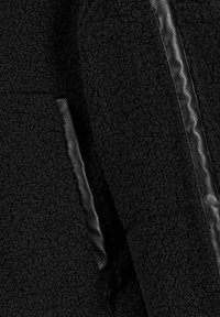Schwarzer Fleece-Mantel aus strukturiertem Stoff, mit einer glatten Lederakzent entlang der Naht. Verfügt über eine seitliche Tasche für zusätzlichen Komfort.