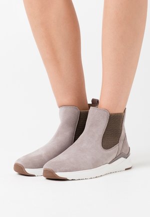 Bottes basses gris clair avec une finition en daim lisse, des panneaux latéraux en mesh, et une semelle en caoutchouc blanche avec un accent marron le long de la base.