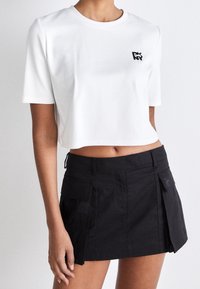 Bílá zkrácená tričko s krátkým rukávem a černým logem, spárované s černou sukní ve stylu kilt s kapsami a detailním klopovým zapínáním.