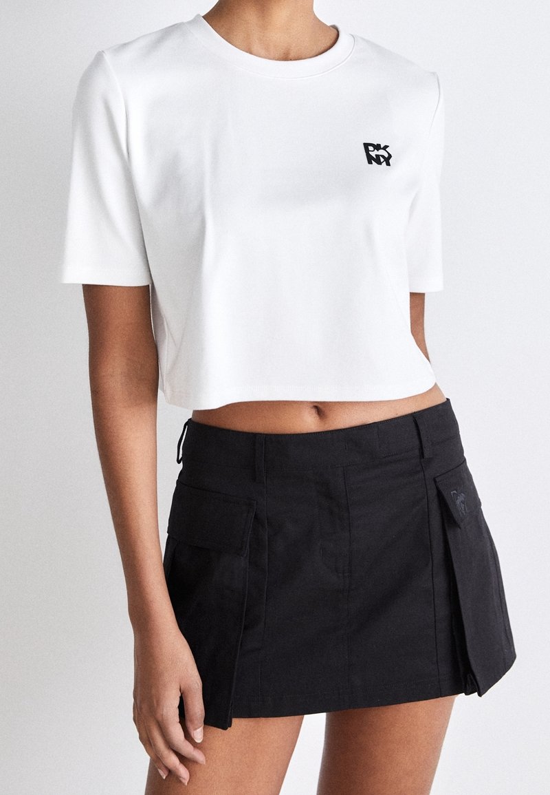 Bílá zkrácená tričko s krátkým rukávem a černým logem, spárované s černou sukní ve stylu kilt s kapsami a detailním klopovým zapínáním.