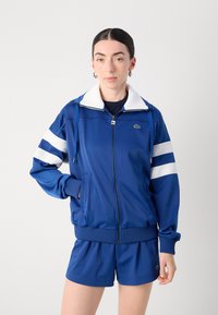 Casaco atlético azul com fecho éclair, com riscas e colarinho brancos, combinado com calções azuis a condizer. Apresenta punhos canelados e detalhe do logótipo.