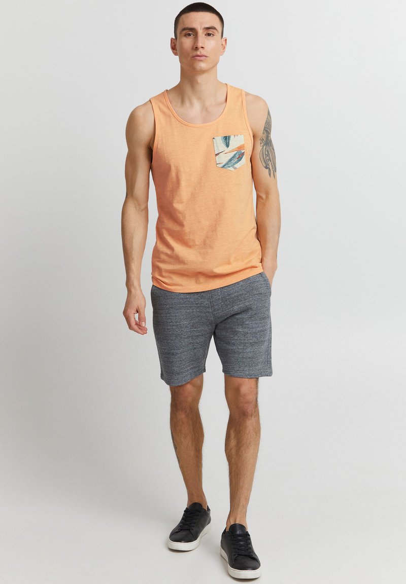 Tricou portocaliu fără mâneci cu o buzănele cu model. Pantaloni scurți gri cu o textură netedă. Adidași negri cu o talpă albă. Design simplu și casual.