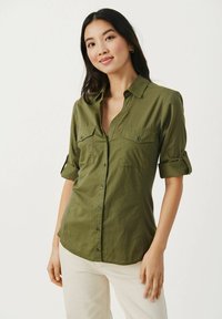Camicia verde oliva con bottoni, maniche arrotolate, due taschini sul petto e tessuto testurizzato. Abbinata a pantaloni chiari.