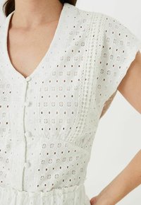 Biała sukienka z ażurowej tkaniny, z dekoltem w kształcie litery V, krótkimi rękawami, zapięciami na guziki oraz haftowanym zdobieniem wzdłuż bocznych szwów.