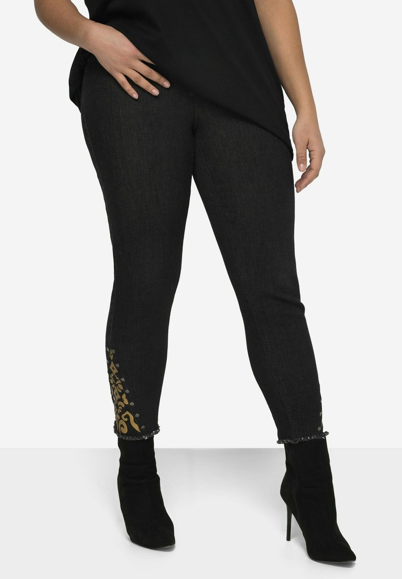 Jeans de mezclilla negros con un diseño ajustado, dobladillo adornado con patrones dorados y tachuelas, combinados con botas de tacón alto negras.