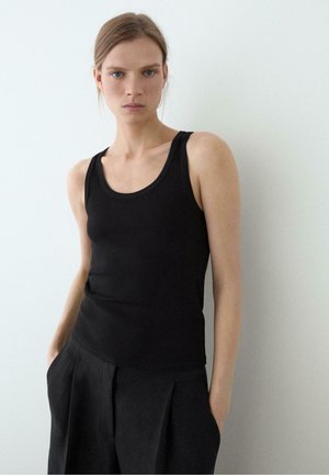 Jeune femme à la peau claire et aux cheveux tirés en arrière, portant un haut noir sans manches et un pantalon noir, debout devant un fond blanc uni.