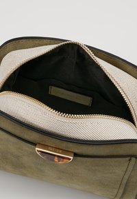 Interno della borsa in suede verde con zip dorata, caratterizzato da una fodera in rete beige e una tasca più piccola con etichetta nera.