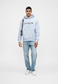 Svetlá modrá mikina s kapucňou a predným vreckom, s šnúrkami na zaviazanie, zdobená logom "Calvin Klein". V páre so svetlomodrými džínsami a bielymi teniskami.