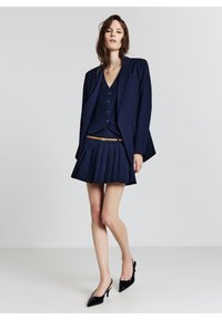 Blazer marine à fermeture à un bouton, top sans manches assorti et jupe mini plissée. Des escarpins en vernis noir complètent la tenue.