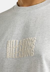 Sudadera de algodón gris con el texto en relieve "BILLABONG" en crema. Cuello redondeado y textura suave. El diseño presenta un efecto de letras superpuestas.
