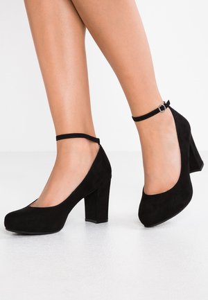 Mustat mokkanahkaiset korkokengät, joissa on pyöreä varvas, paksu korko ja nilkkahihna, jossa on pyöreä metallikoriste. Sileä pinta, tyylikäs muotoilu.