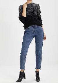 Frau trägt einen schwarz-weiß gesprenkelten Pullover, hoch taillierte blaue Jeans mit ausgefransten Säumen und schwarze Leder-Stiefeletten mit Absatz.