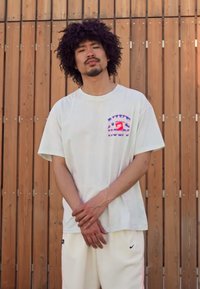 Hvid bomulds t-shirt med et multicoloreret logo-patch på venstre bryst, parret med hvide shorts. Baggrunden består af træpaneler.